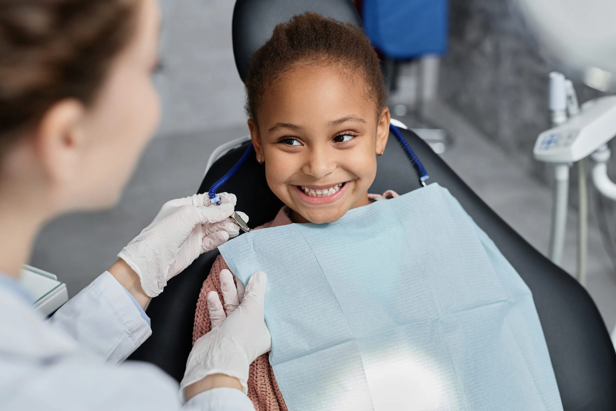 Young girl smiling in dental chair while dentist in gloves places bib on her
