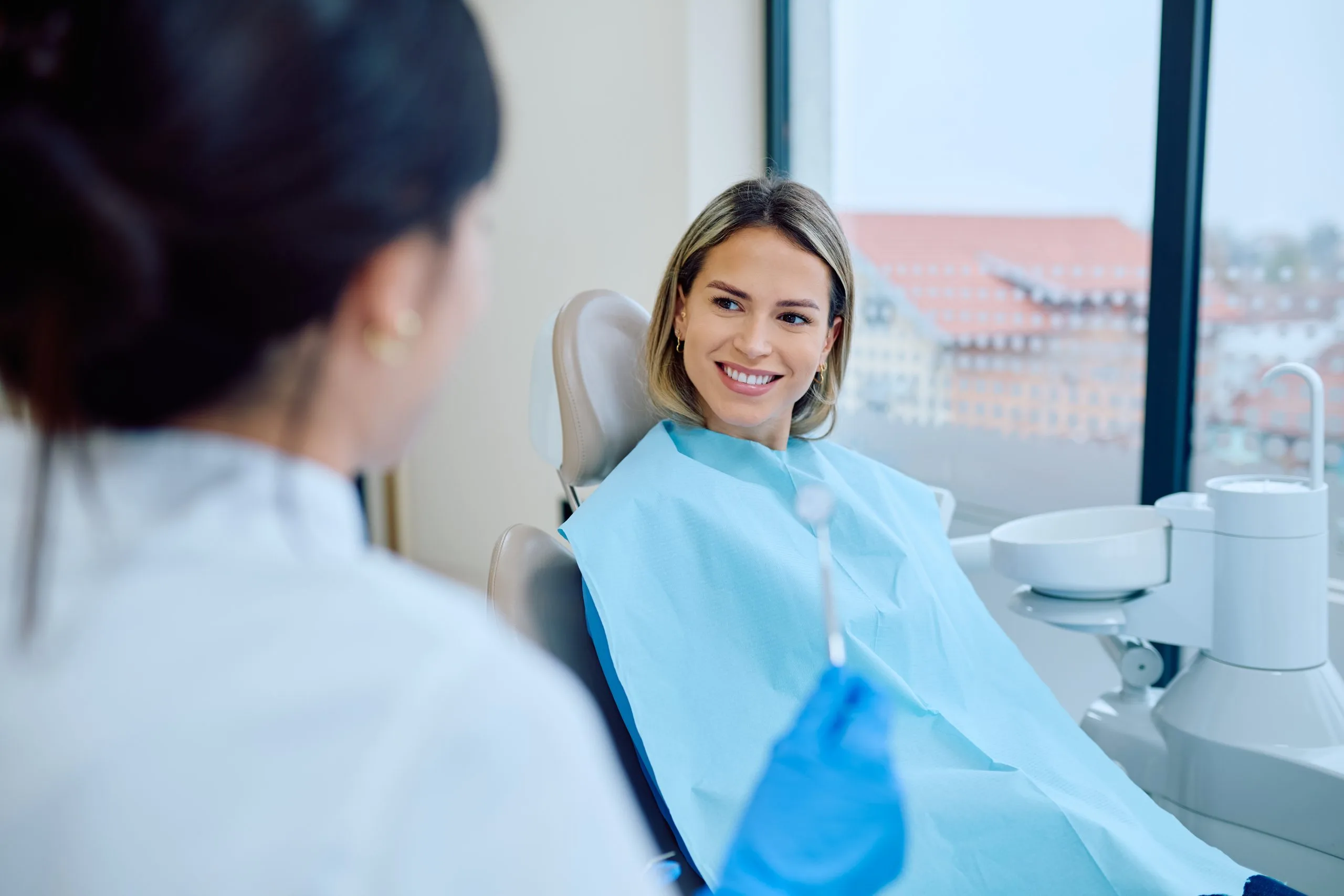 Smiling patient in blue bib sits in dental chair facing dentist during consultation