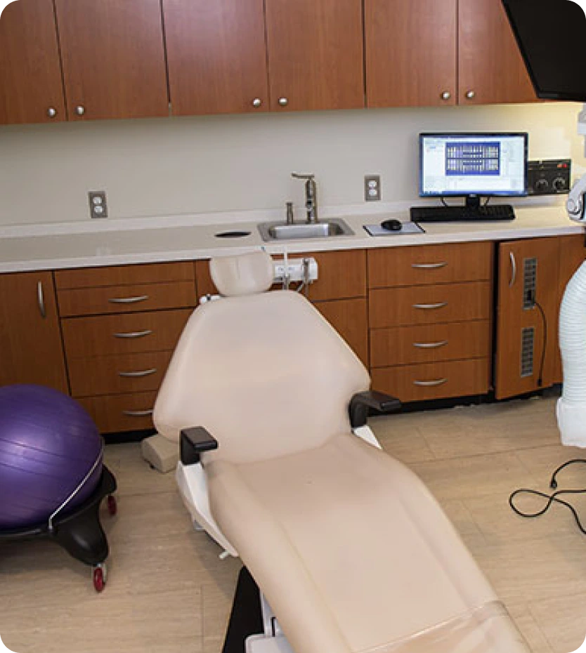 Dental treatment room with cream-colored chair, wood cabinetry, computer monitor, and exercise ball